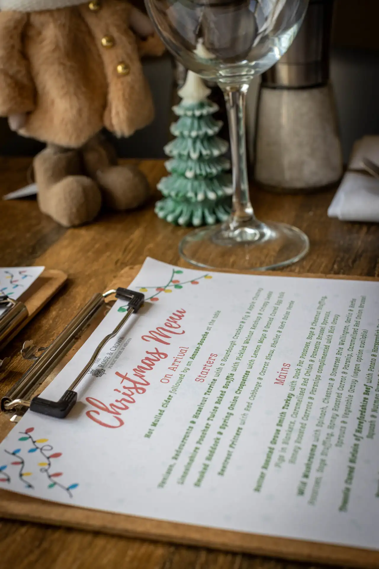 The Rose & Crown Christmas menu on a clipboard, laid on a wooden table with wine glasses and little Christmas trees